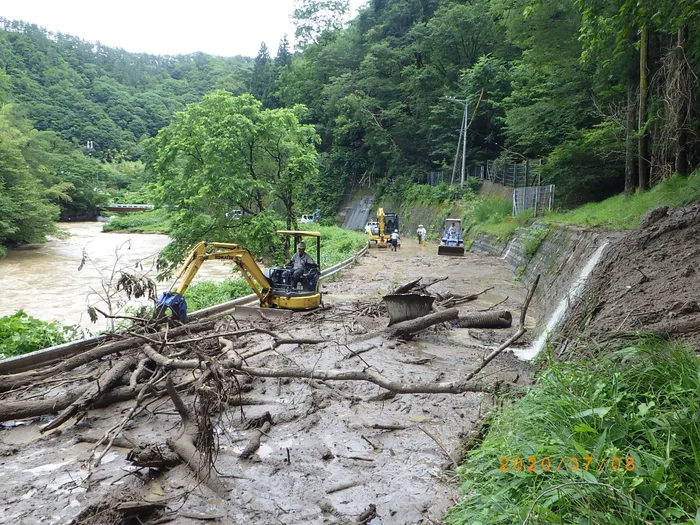 土砂と流木に埋まった川沿いの道路で、復旧にあたる小型のバックホウ