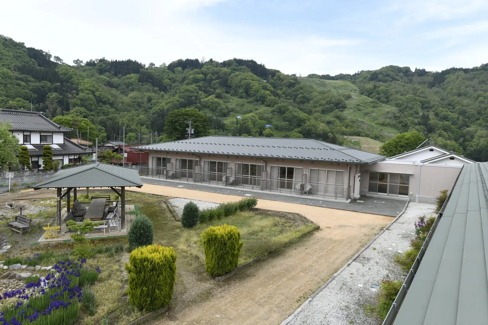 山を背に建つ平屋の施設。手前の庭に東屋と植栽が並ぶ